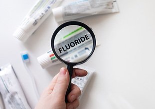 Hand holding magnifying glass over toothpaste tube displaying “FLUORIDE”
