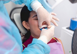 Young woman in dental chair closing eyes as gloves hands place fluoride trays over her teeth