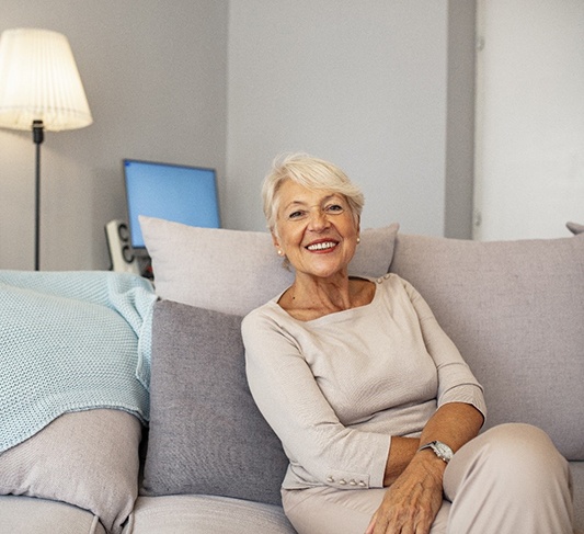 An older woman sits comfortably on her couch and smiles after receiving dental implants