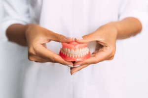 A person wearing white in the background holding a set of dentures with both hands in the foreground